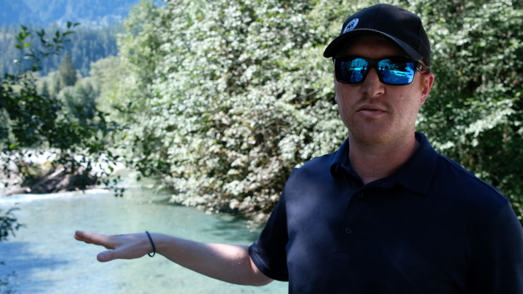 A male biologist wearing sunglasses is on a bridge pointing towards a creek full of spawning salmon