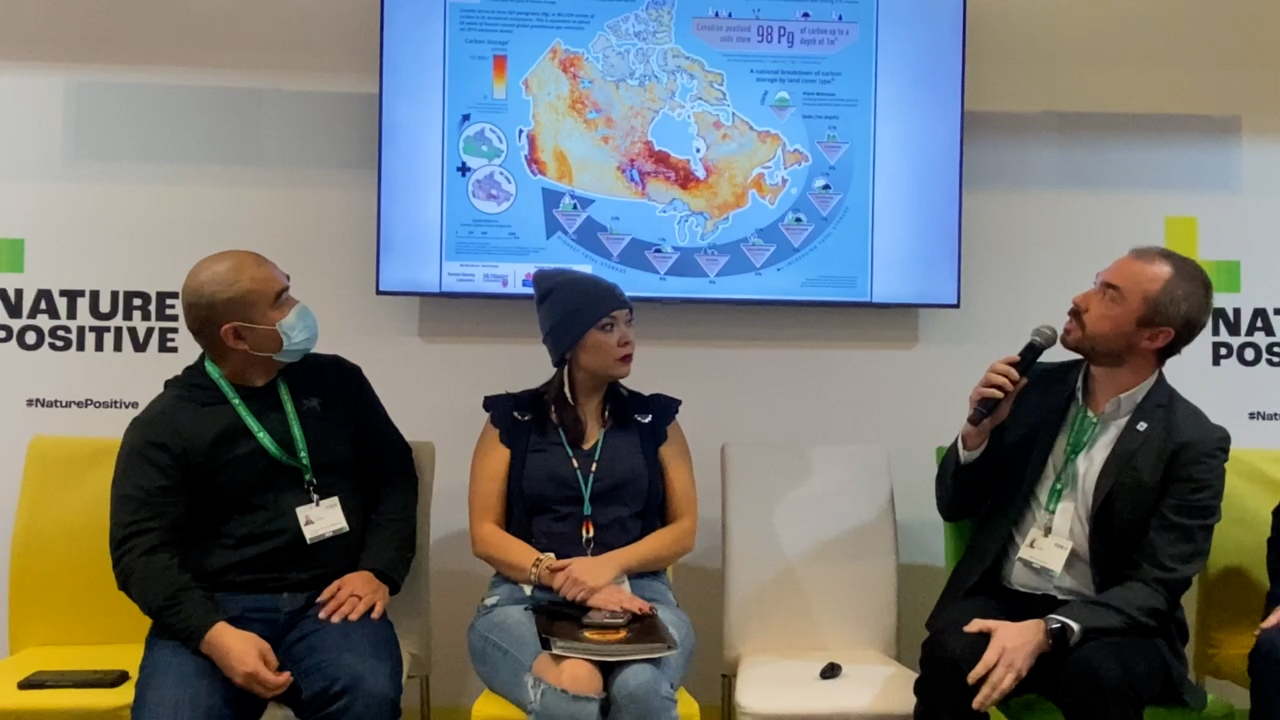 A woman sitting between two men in front of a map showing Canada’s carbon stocks)