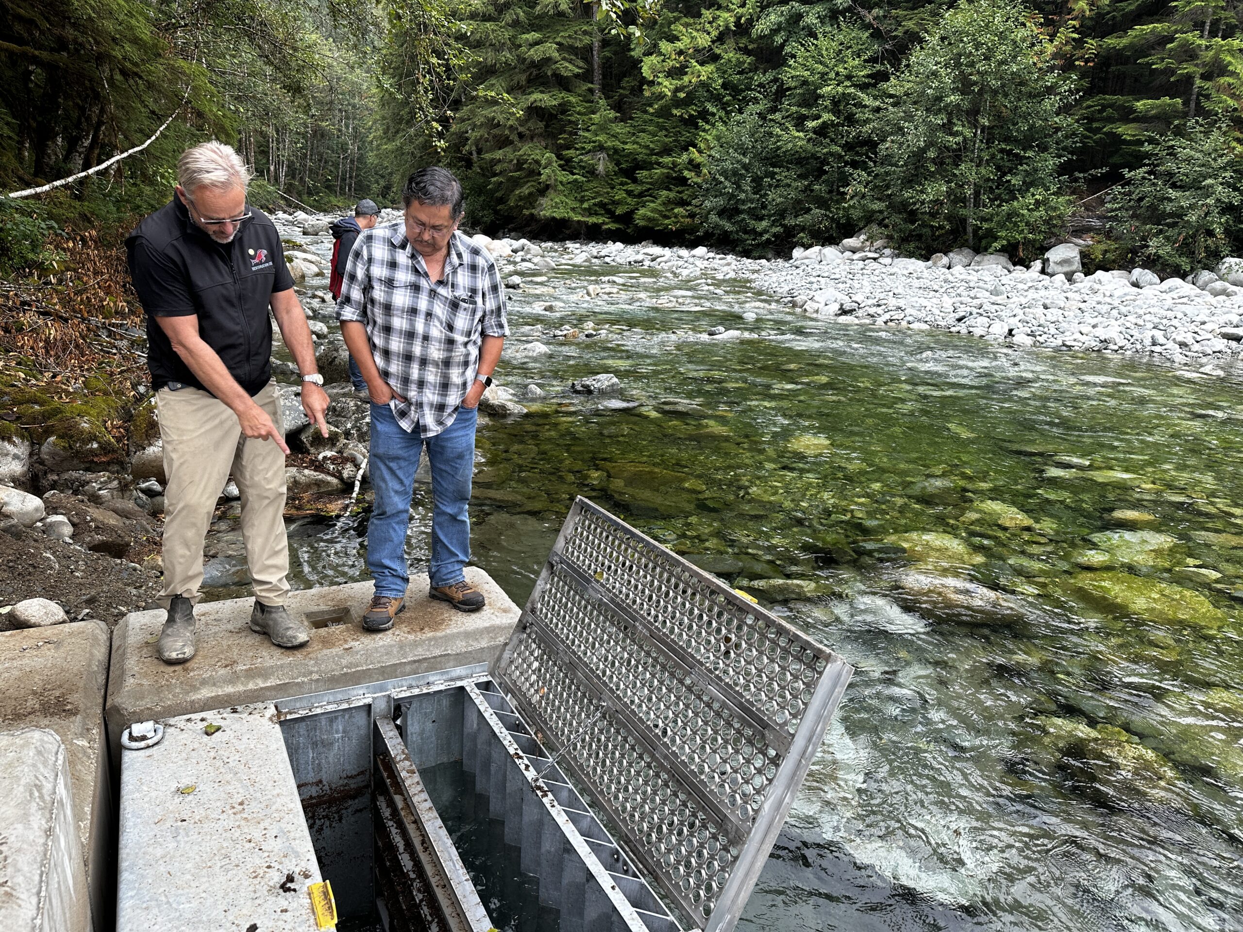 Two men in a forest looking at an intake valve beside a creek