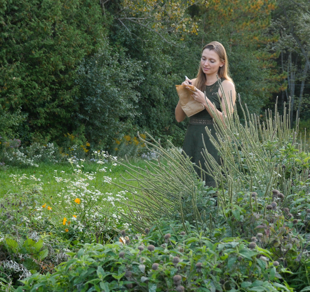 A person stands beside a flourishing garden, writing on a paper bag that holds seeds.