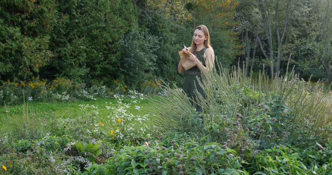 A person stands beside a flourishing garden, writing on a paper bag that holds seeds. 