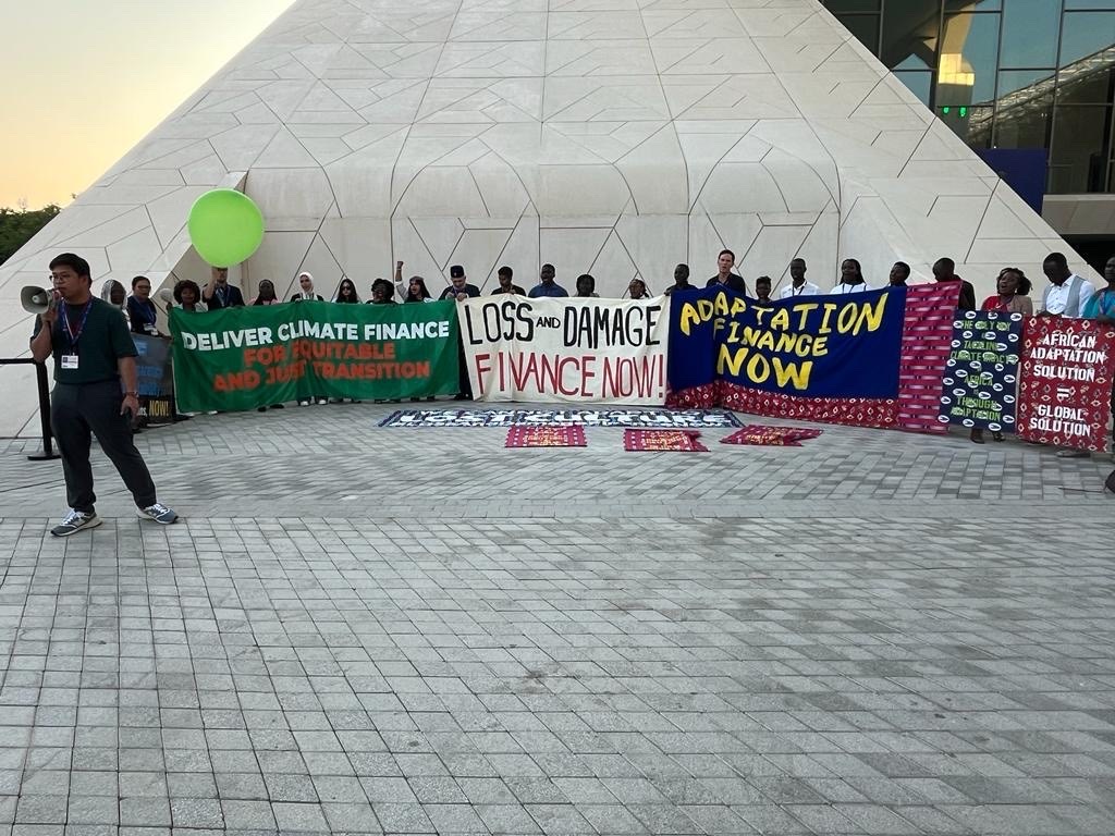 A group of people holding signs that say "deliver climate finance," "loss and damage finance now," and "adaptation finance now"