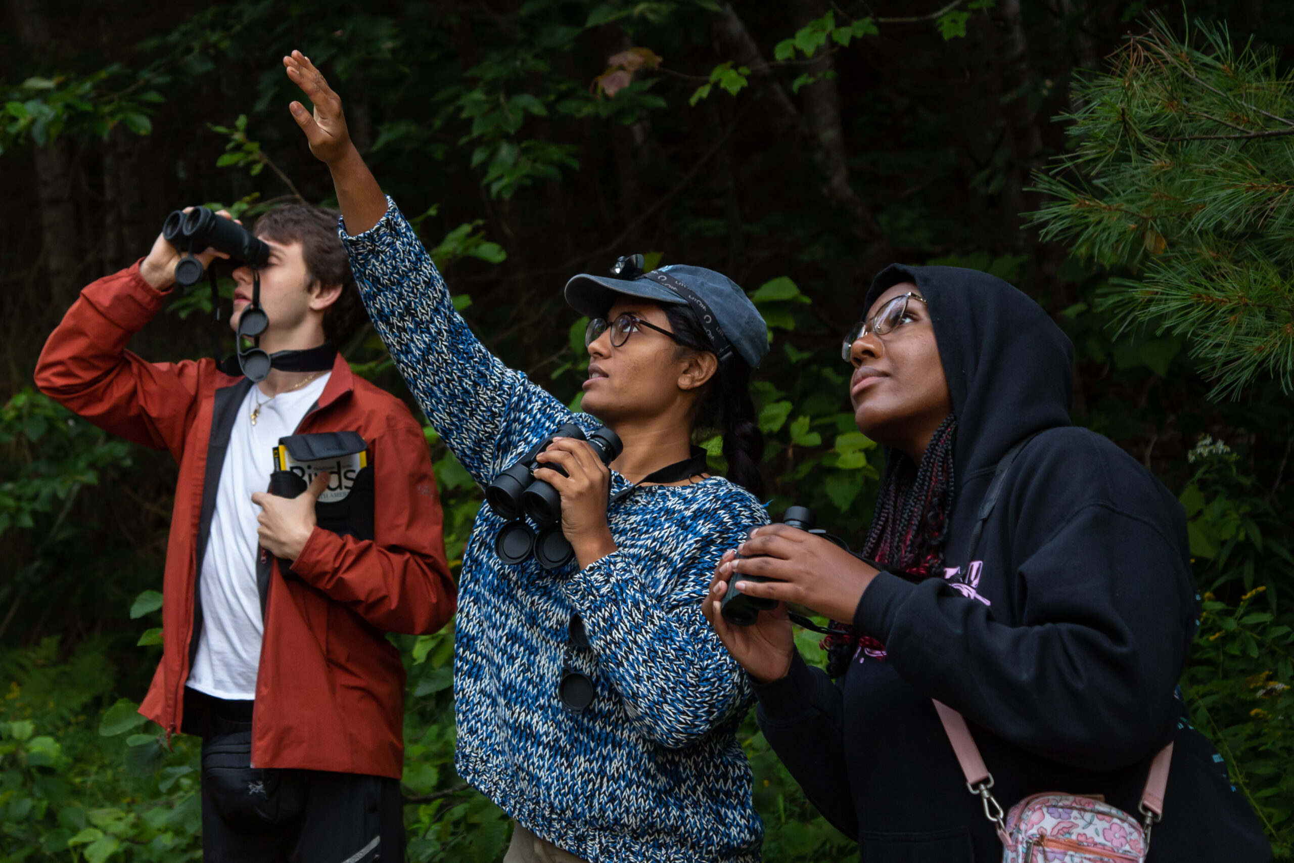 Young woman, with a male and female student on either side of her, holds binoculars in one hand and points toward the tree canopy