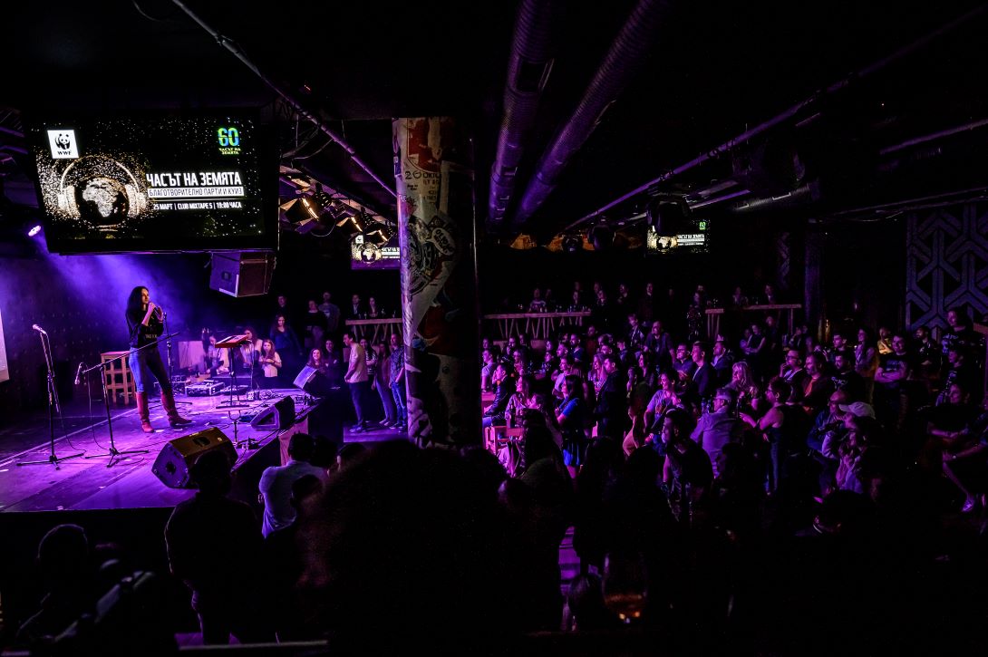 A person onstage, standing below a sign showing the WWF and Earth Hour logos, addressing a room full of people.