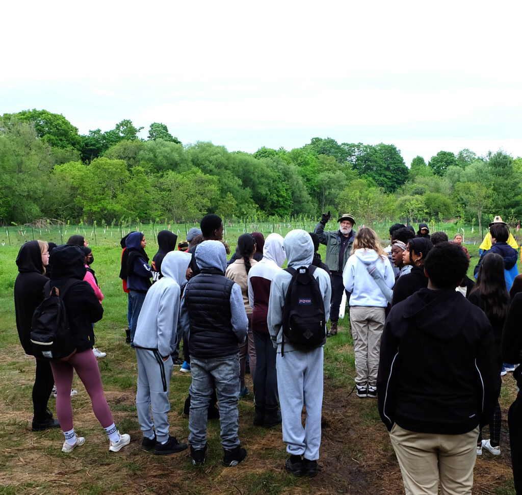 A man talking to a group of young students in front of small newly planted trees. 