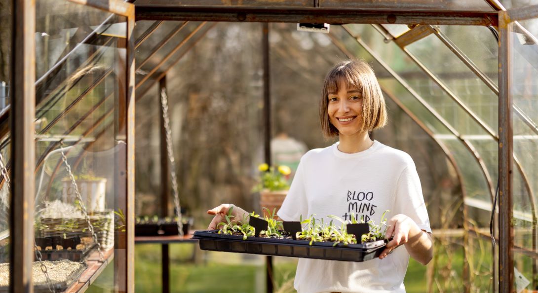 A smiling young adult carries a tray of seedlings from a small greenhouse into a yard on a sunny morning.