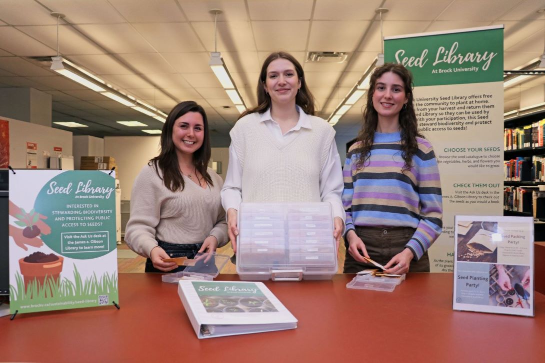 Three people holding containers of seed envelopes smile standing at a counter in a library beside three signs promoting the seed library.