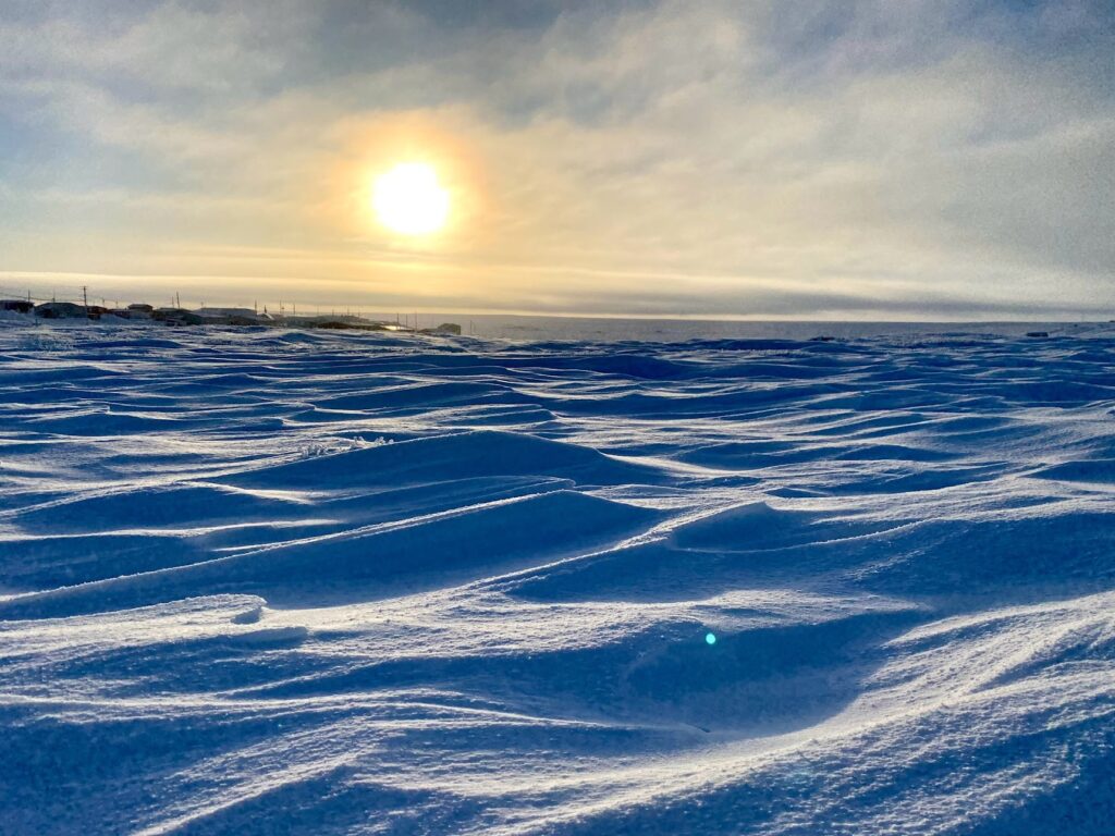 Scène de coucher de soleil montrant des ondulations de neige balayées par le vent au premier plan, avec des bâtiments et l’océan au loin